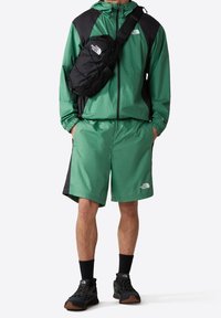 Veste en nylon verte avec des accents noirs, shorts assortis et baskets noires. Comprend un sac bandoulière noir et dispose de poignets élastiques et de détails de logo.