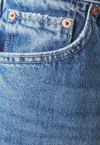 Ljusblå denimtyg med en blekt textur. Har en framficka med kopparnitar och synliga sömdetaljer.