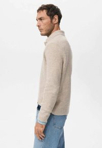 Beżowy sweter z dzianiny z kołnierzem, ściągaczami na mankietach i dole oraz miękką fakturą. Noszony z jasnoniebieskimi dżinsami.