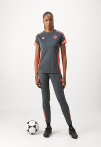 Camiseta deportiva gris con acentos naranjas, combinada con leggings grises a juego. Presenta el logo de Adidas, un balón de fútbol y botas negras.