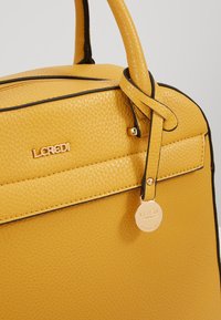 Żółta torebka skórzana o fakturze z czarnymi akcentami, o zaokrąglonym kształcie, z górnymi rączkami i metalową etykietą z logo.