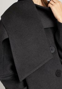 Abrigo de lana negro con un gran cuello drapeado, diseño de doble botonadura y botones negros. La tela parece suave con un acabado texturizado.