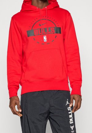 Mand iført rød Chicago Bulls hættetrøje med sort basketballgrafik og sorte bukser med hvid Bulls-logo og tekst på låret.