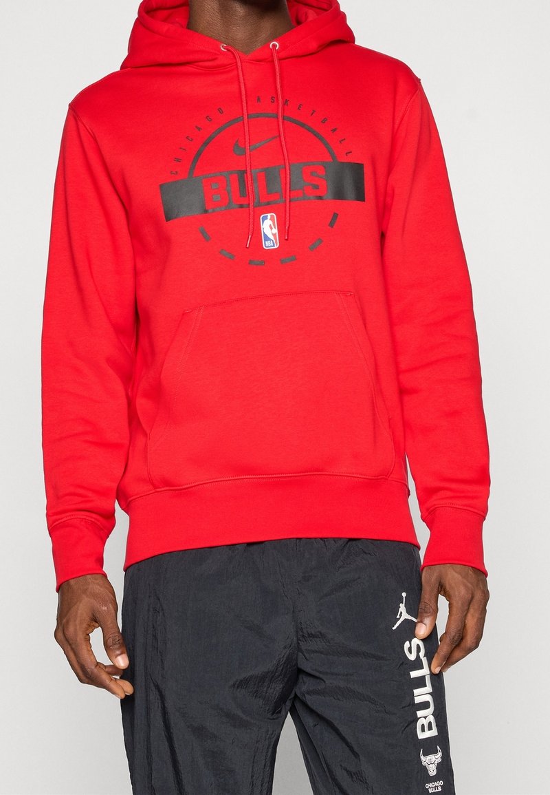 Homme portant un sweat à capuche rouge Chicago Bulls avec un graphique de basket-ball noir et un pantalon noir avec le logo blanc des Bulls et du texte sur la cuisse.