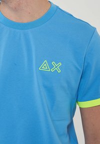 Maglietta in cotone blu con logo geometrico giallo sul petto e dettagli gialli vivaci sulle maniche. Tessuto morbido, scollo rotondo.