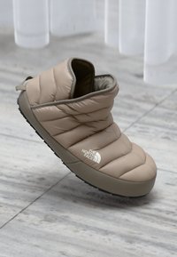 Chausson beige matelassé avec une texture douce, semelle en caoutchouc et tirant à l'arrière. Présente un logo sur le côté et une silhouette arrondie.