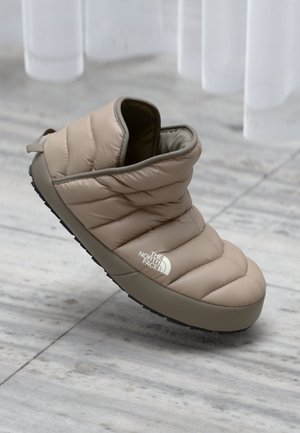 Beige quilted slipper with a soft texture, rubber sole, and rear pull tab. Features a logo on the side and a rounded silhouette.