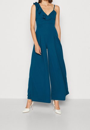 Teal-huonepuku yhdellä olkaimella, joissa on volangi, kapea yläosa, leveälahkeinen muotoilu ja sileä pinta. Yhdistetty valkoisiin korkokenkiin.