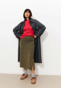 Manteau à carreaux gris sur un pull rouge, associé à une jupe olive côtelée avec des boutons sur le côté, des chaussettes grises et des chaussures beiges à lacets.