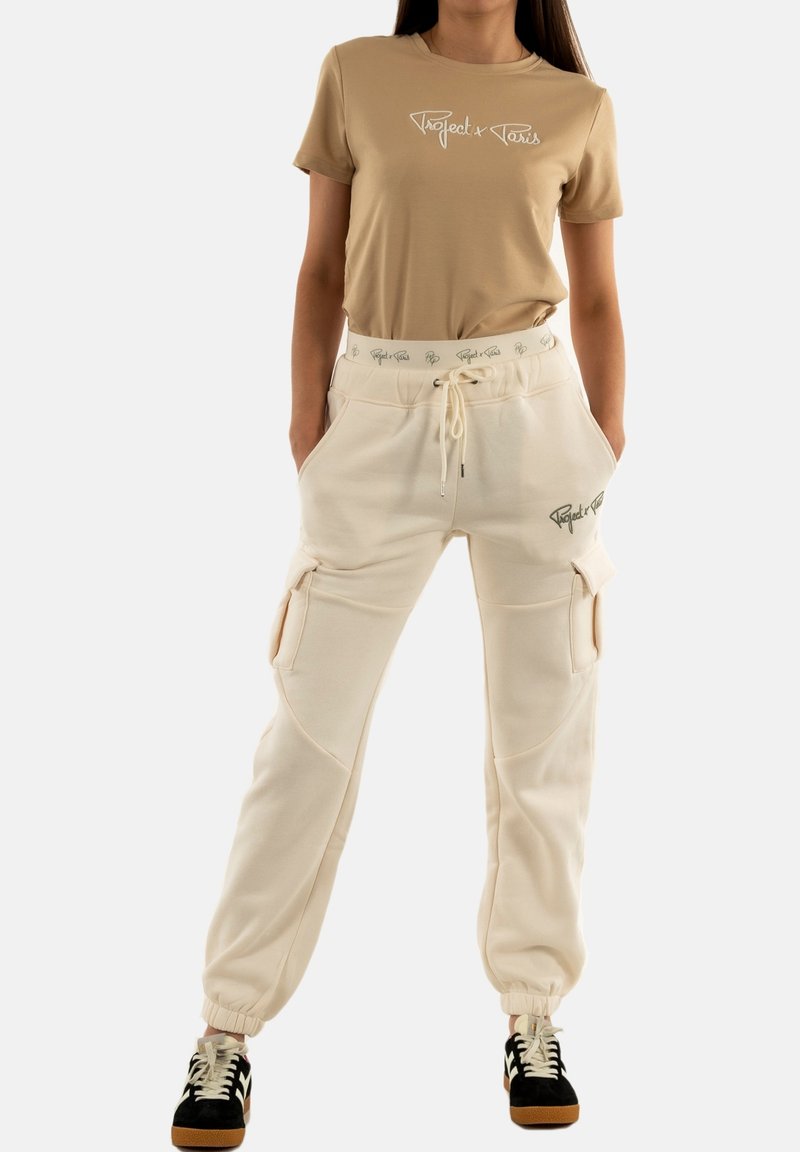 Camiseta beige con logo blanco, combinada con pantalones cargo crema que tienen bolsillos laterales y puños elásticos. Zapatillas negro y blanco completan el conjunto.