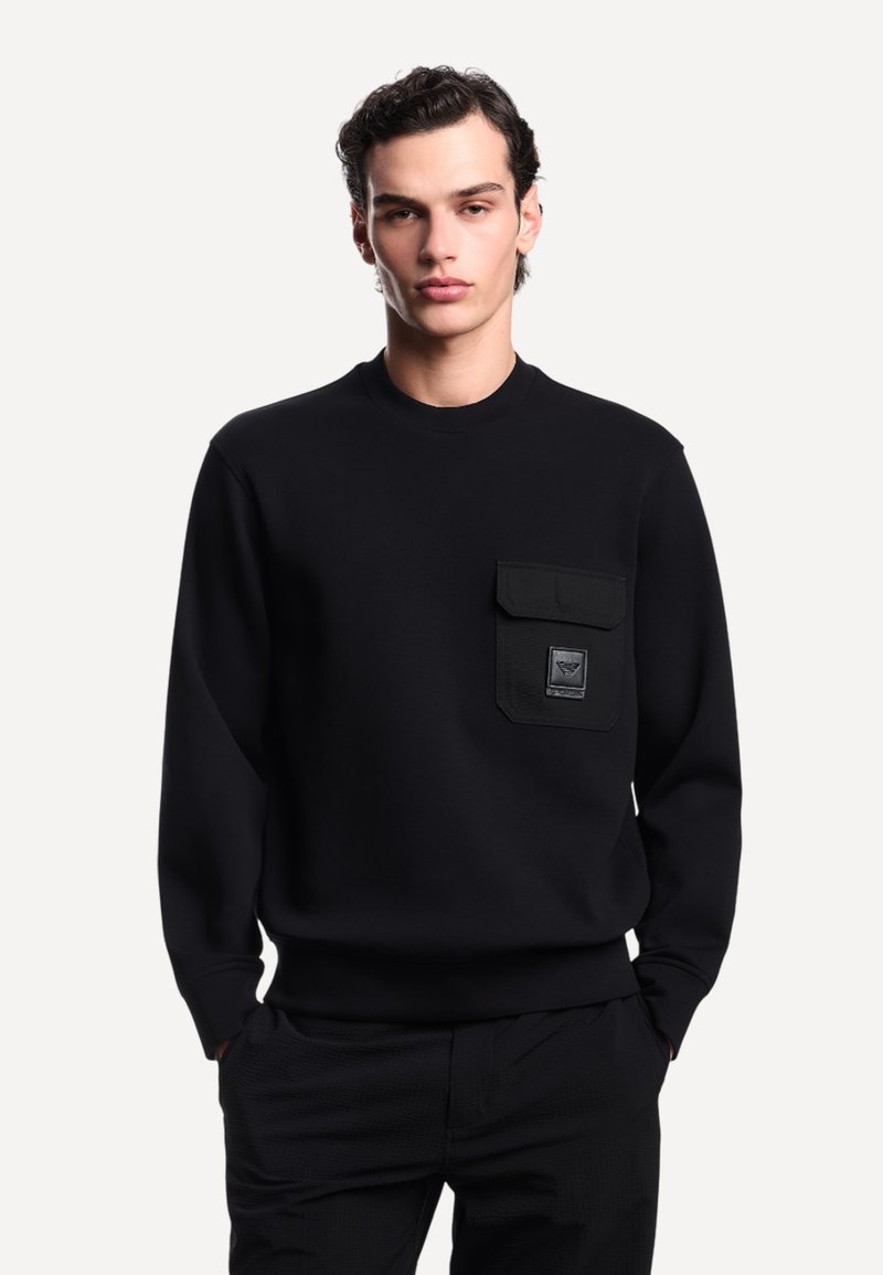 Giovane uomo che indossa un felpa nera con tasca a pattina e un piccolo logo sul lato sinistro del petto, con le mani nelle tasche dei pantaloni neri, su uno sfondo neutro.