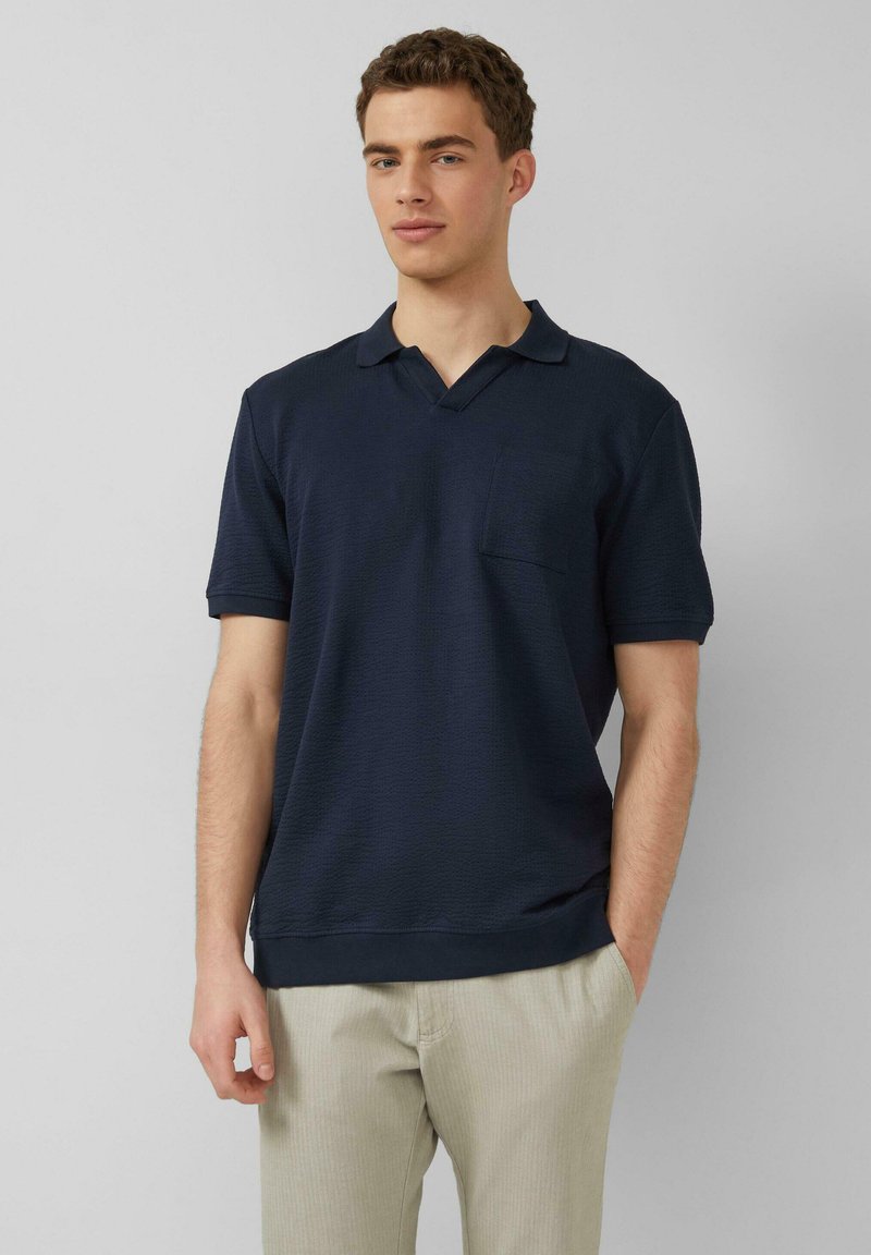 Junger Mann mit kurzen lockigen Haaren, der ein marineblaues Polohemd mit kurzen Ärmeln und hellbeige Hosen trägt und vor einem einfarbigen grauen Hintergrund steht.