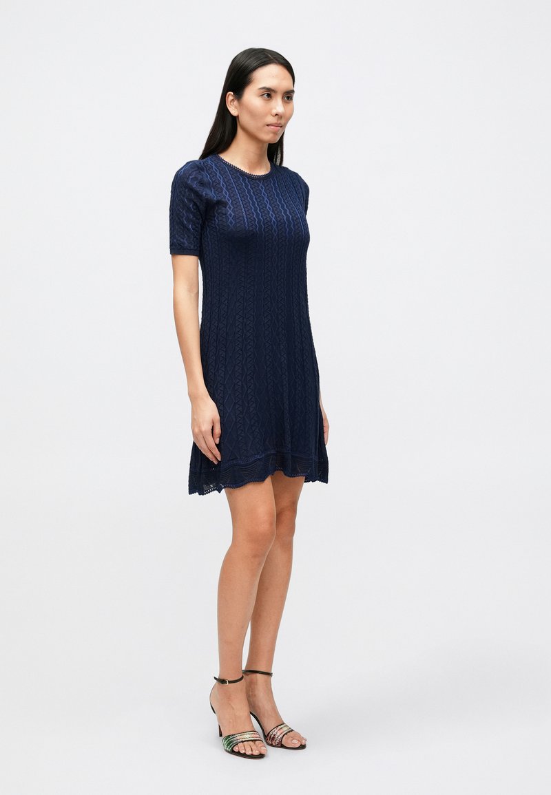 Navy blue knitted dress with short sleeves, featuring a textured pattern and scalloped hem. Paired with multicolored strappy sandals.