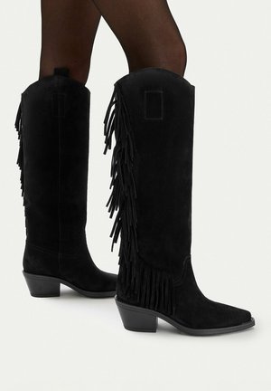 Schwarze kniehohe Stiefel aus Wildleder mit seitlichen Fransen, getragen zu transparenten schwarzen Strumpfhosen vor weißem Hintergrund.