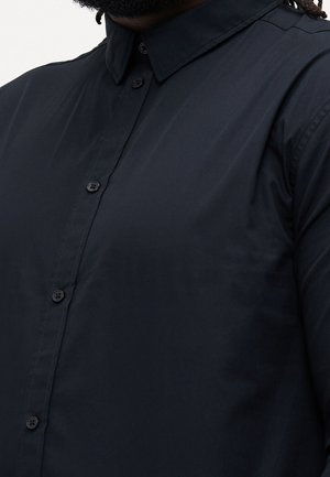 Camicia blu navy scuro con bottoni e colletto indossata da una persona con la pelle scura e capelli neri ricci, in primo piano.