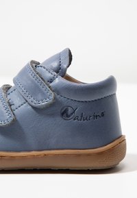 Modrá kožená bota s dvěma suchými zipy, texturovaným povrchem, kulatou špičkou a hnědou gumovou podrážkou. Na straně se nachází vyražené logo "Naturino".