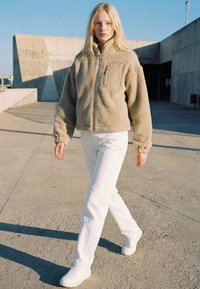 Młoda kobieta spacerująca na zewnątrz, ubrana w beżową polarową kurtkę, białe spodnie i białe trampki na tle betonowych konstrukcji i czystego nieba.