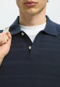 Námořnická modrá polo tričko s texturovanými vzory, tříknoflíkovou légou a kontrastním bílým tričkem pod ním. Ruka držící popruh tašky.