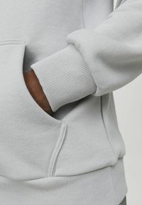Ljust grå sweatshirt med ribbade muddar och fickdetaljer. Materialet verkar mjukt med en slät yta och sitter tajt vid handleden.