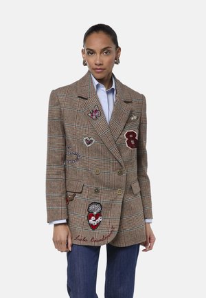 Mujer con blazer cruzado de cuadros con parches coloridos, camisa azul clara y vaqueros azul oscuro sobre un fondo liso.