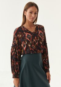 Patterned blouse with a v-neckline and long puff sleeves. Colors include dark hues with floral and geometric designs. Worn with a green skirt.