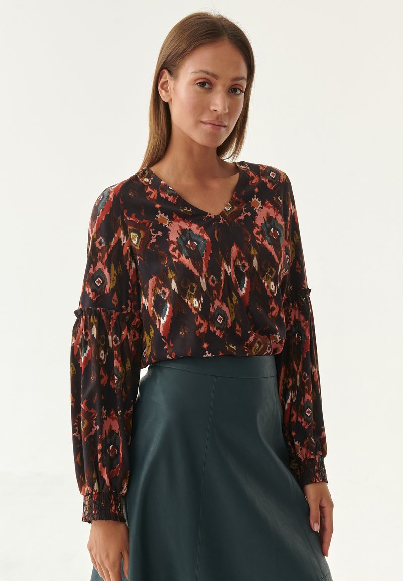 Patterned blouse with a v-neckline and long puff sleeves. Colors include dark hues with floral and geometric designs. Worn with a green skirt.