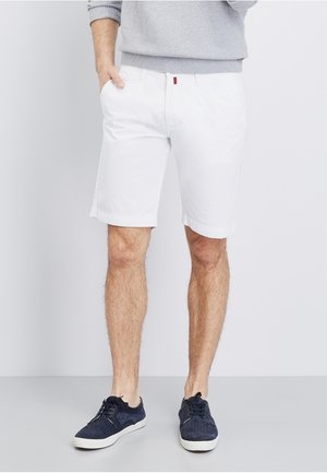 Mand iført hvide shorts til knæet, grå sweater og mørkeblå casual sneakers, stående med den ene hånd i lommen.