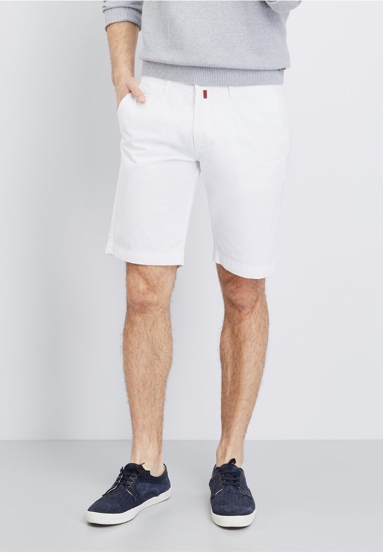 Homme portant un short blanc jusqu'aux genoux, un pull gris et des baskets décontractées bleu foncé, debout avec une main dans la poche.