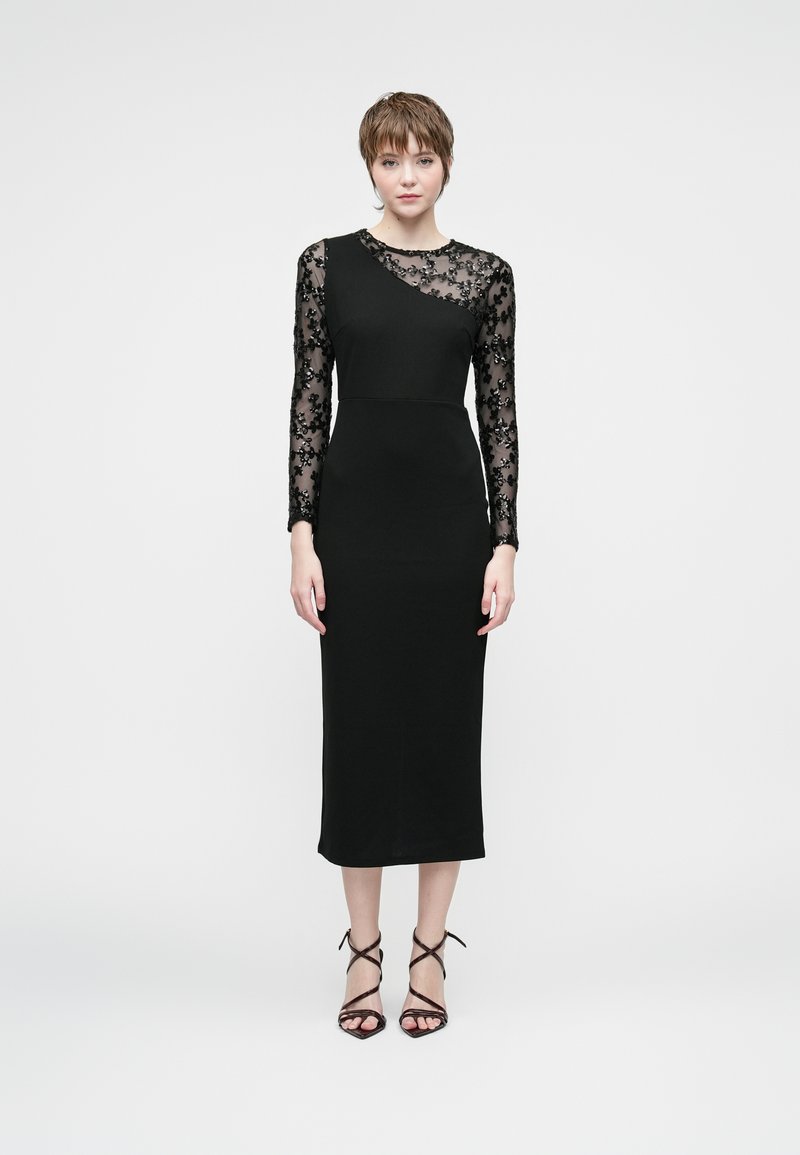 Naine seisab mustas asümmeetrilises kleidis, millel on läbipaistvad lillemustriga pikad varrukad ja mustad rihmadega kontsad, tavalise valge taustaga.