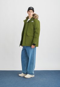 Πράσινο μπουφάν με επένδυση και κουκούλα με γούνα, διαθέτοντας πολλές τσέπες και φερμουάρ. Συνδυασμένο με φαρδιά μπλε τζιν και αθλητικά παπούτσια.