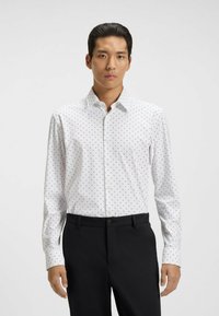 Camicia bianca con piccoli motivi rossi e blu, maniche lunghe, colletto classico, abbinata a pantaloni neri testurizzati.