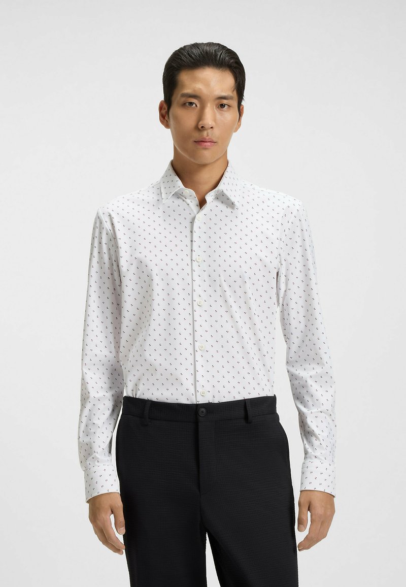 Camicia bianca con piccoli motivi rossi e blu, maniche lunghe, colletto classico, abbinata a pantaloni neri testurizzati.