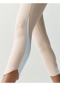 Béžové legíny se světle modrým bočním panelem, hladká textura, přiléhavý střih a prošívané detaily na okrajích a lemech panelu.