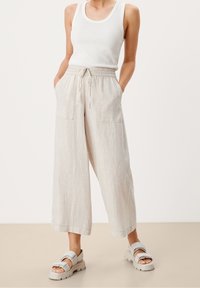 Licht beige linnen wijde broek met een trekkoord rond de taille, gestructureerde stof en twee voorzakken. Gepaard met witte platte sandalen.