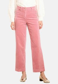 Žena oblečená v ružových kordovych nohaviciach s vysokým pásom a bočnými gombíkmi, so svetlými nízkymi podpätkami, stojí na bielom pozadí.