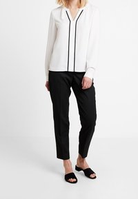 Nainen pukeutuneena valkoiseen pitkähihaiseen paitaan, jossa on mustia yksityiskohtia, mustiin muotonsa mukaiseen nilkkapaitoon ja mustiin avokäsineisiin, seisoo valkoista taustaa vasten.