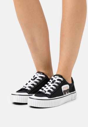 Zwarte lage sneakers met witte veters en dikke zolen, met een klein grafisch patch aan de zijkant, gedragen op blote voeten tegen een witte achtergrond.
