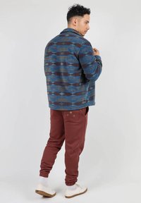 Veste à col bleue avec des motifs abstraits marron et beige, associée à un pantalon de survêtement bordeaux doté d'une poche avec des accents décoratifs.