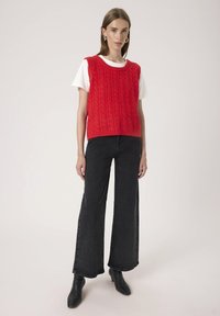 Woman wearing a red cable-knit sleeveless sweater over a white t-shirt, black wide-leg jeans, and black heeled boots, standing against plain background.