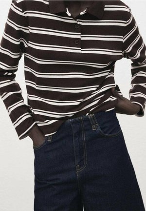 Persoon draagt een bruin-wit gestreept poloshirt met lange mouwen, ingestopt in een high waist donkerblauwe spijkerbroek met handen in de zakken.