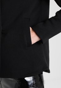 Sort langærmet jakke med en sidelomme, lavet af et glat, strækbart stof. Teksturen virker blød, og designet er minimalistisk.