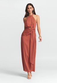 Moteris vilkinti rudą halterio tipo maksi suknelę su šonine juostele, šlepetes su geltonais karoliukais ir auksinę papuošalų kolekciją ant baltos fono.