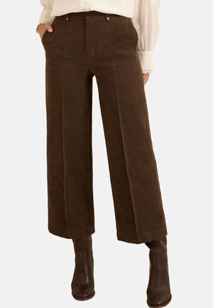Pantaloni maro cu picior larg și cusături frontale, purtați cu ghete maro asortate și o bluză albă cu mâneci lungi, parțial băgată în pantaloni.