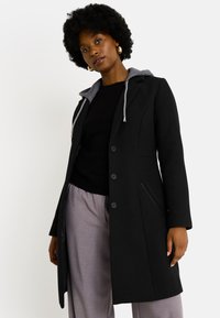 Abrigo negro con capucha gris, diseño estructurado, botones frontales, bolsillos laterales y un acabado elegante. Lleva puesto un suéter negro y pantalones claros.