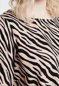 Persona bionda che indossa una blusa beige con strisce zebrate nere e maniche lunghe, mostrando il torso superiore e il décolleté.