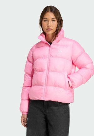 Une jeune femme se tient debout, portant une doudoune rose vif par-dessus un t-shirt noir et un jean foncé, les mains dans les poches de la veste, sur un fond neutre.