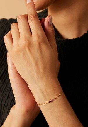 Gouden kettingarmband met een rij kleine rode edelstenen. De armband ligt op een hand met een natuurlijke huidtextuur en licht gemanicuurde nagels.
