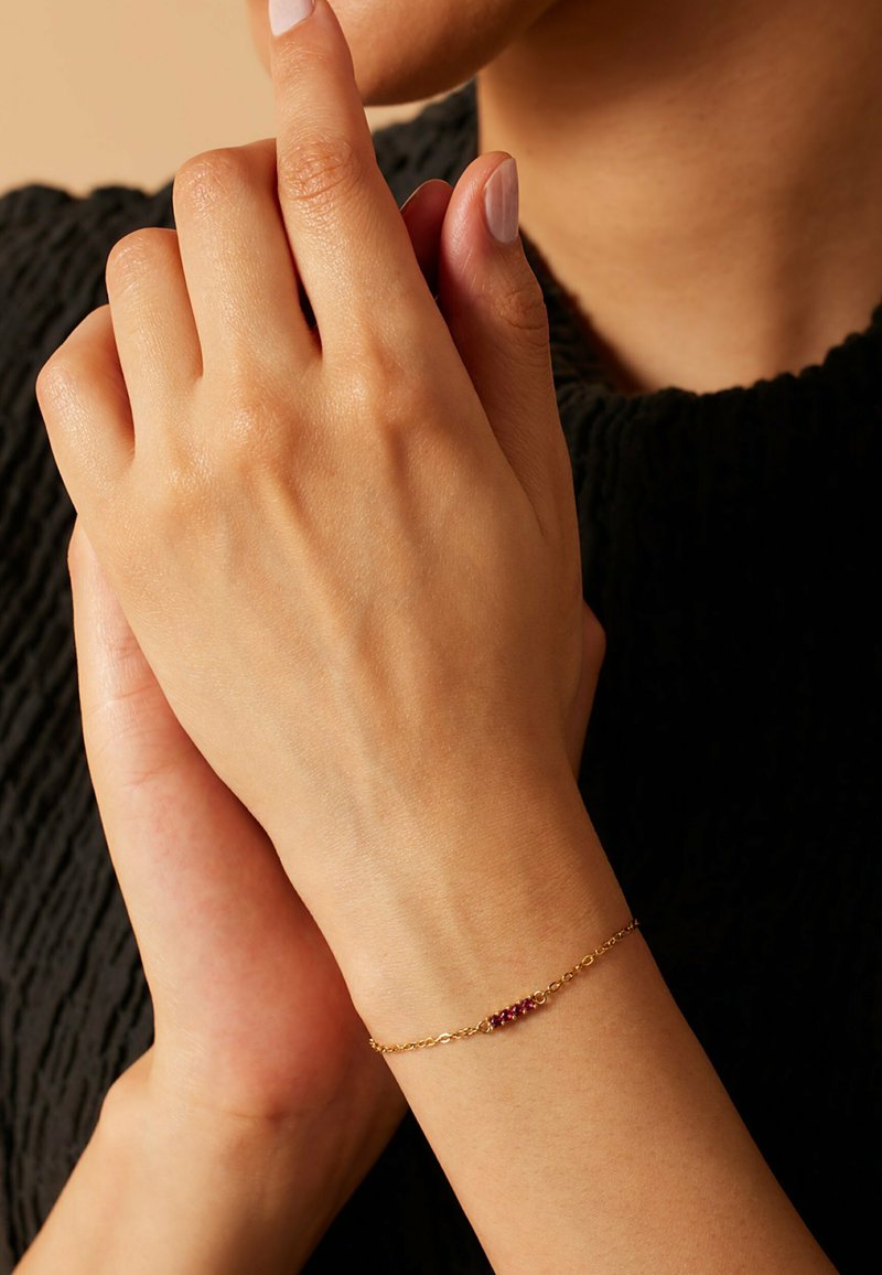 Gouden kettingarmband met een rij kleine rode edelstenen. De armband ligt op een hand met een natuurlijke huidtextuur en licht gemanicuurde nagels.