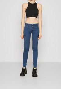 Jeans skinny azul con dos bolsillos laterales y cierre de botón, combinados con un top negro corto y zapatos negros gruesos.