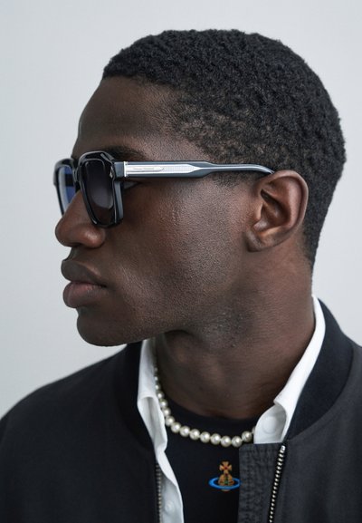 Fekete, téglalap alakú napszemüveg sötét lencsékkel és ezüst díszítésű szárakkal, amelyet egy rövid, texturált hajú személy visel, fekete bombázó dzsekiben.